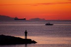 Costa livornese. Una ventina di minuti dopo il tramonto, la Corsica si staglia nettamente sullo sfondo di un cielo arancio vivo. Un pescatore, una barca  che passa ed una nave, sottolineano i vari piani.