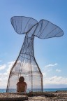 Una persona seduta all'interno della base di una grande scultura in rete metallica che riproduce una coda di balena, con il mare e il cielo azzurro sullo sfondo. L'installazione  un'opera in rete metallica a forma di coda di balena, situata sul molo di Camogli, in Liguria.