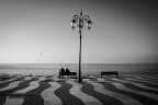 fotografia in bianco e nero che ritrae il lungolago di Lazise, caratterizzato da una pavimentazione a onde, un lampione centrale e due persone sedute su una panchina che guardano il lago.
